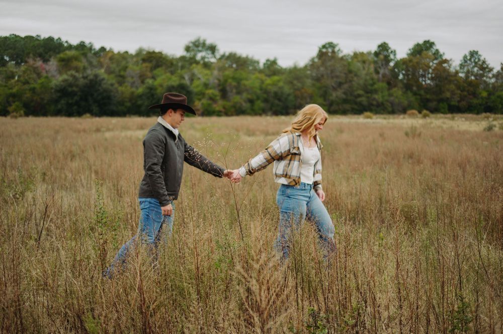 A couple holds hands while walking through a golden autumn field with trees in the background.