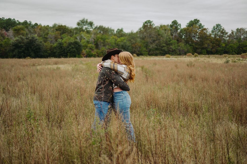 Two people embrace while standing in a golden autumn field with trees in the background.