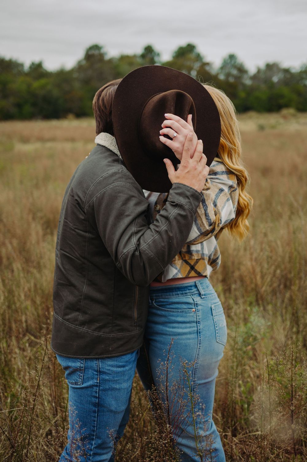 Couple embracing in tall grass field wearing denim and casual attire during outdoor photoshoot.