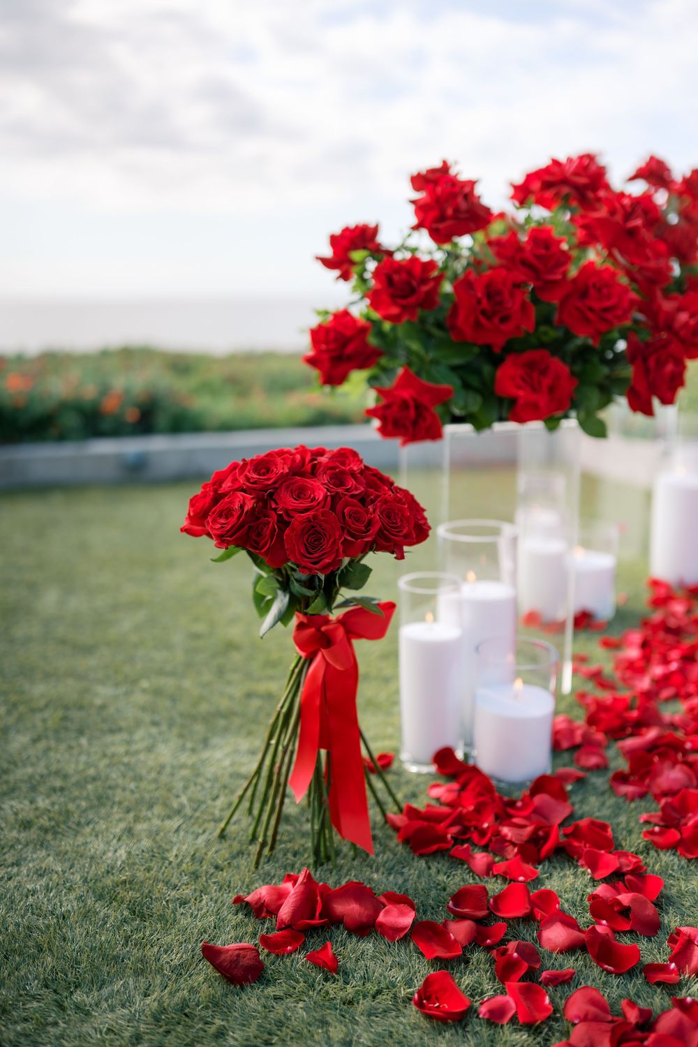 Red rose bouquets with white candles and scattered petals create a romantic outdoor ceremony setting on green grass.