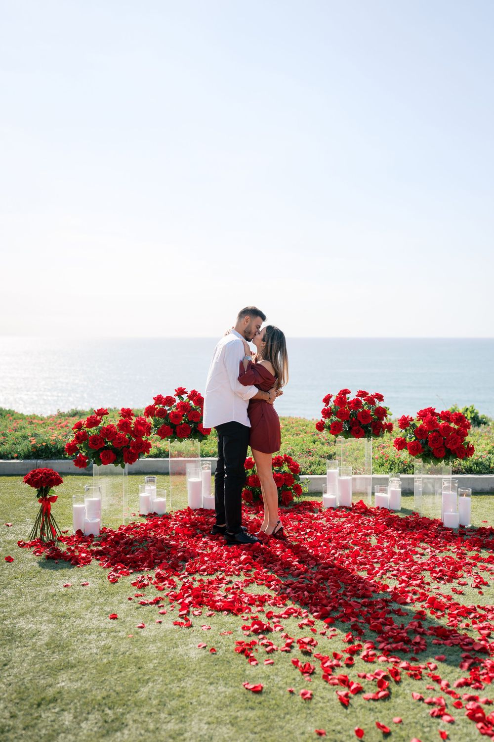 A romantic proposal setup with white candles and red rose petals scattered along a grassy outdoor path.