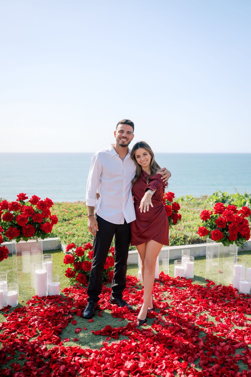 Couple poses together on grass covered in bright red rose petals with ocean view backdrop.