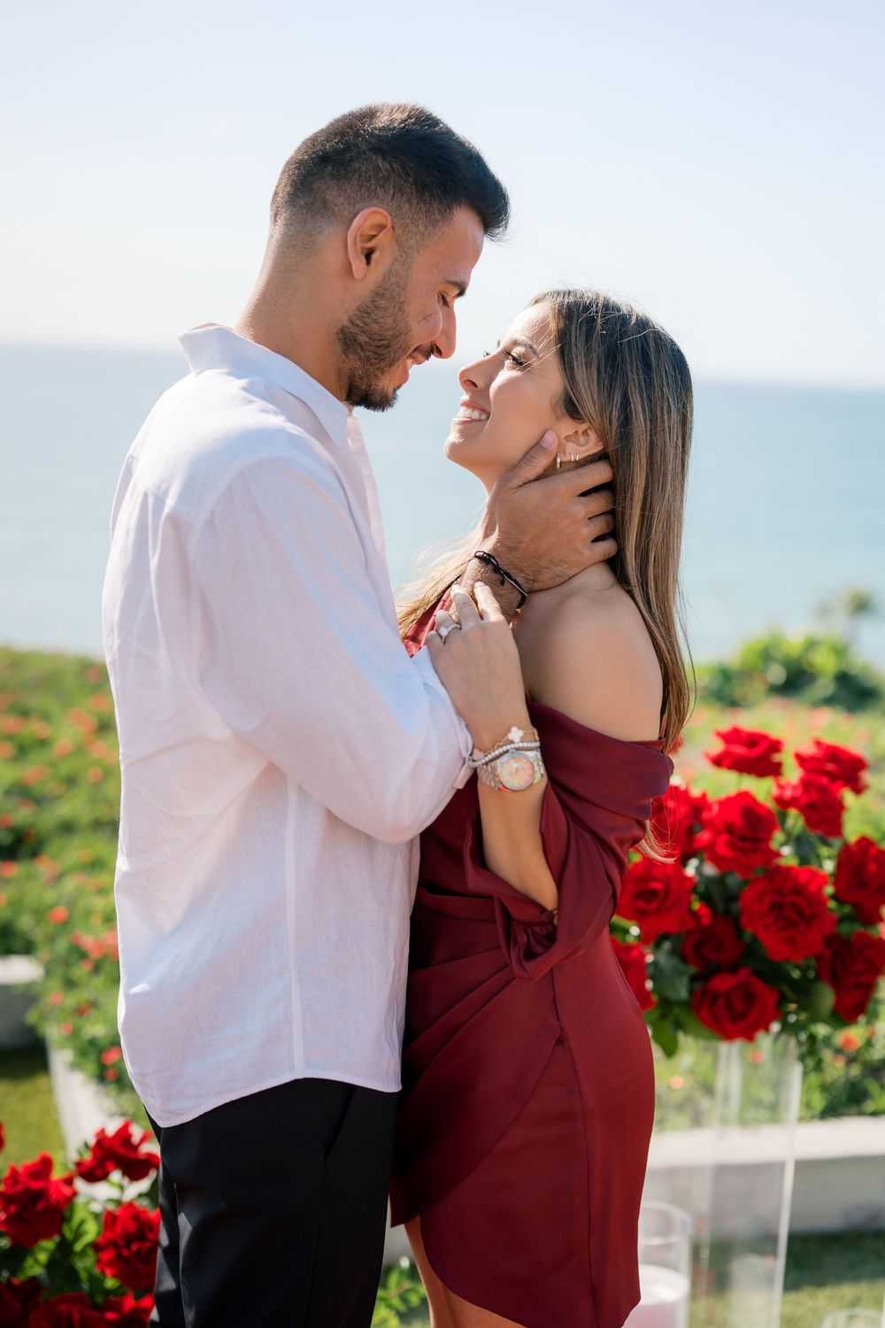 A romantic couple embraces by the ocean with red roses in the foreground on a sunny day.