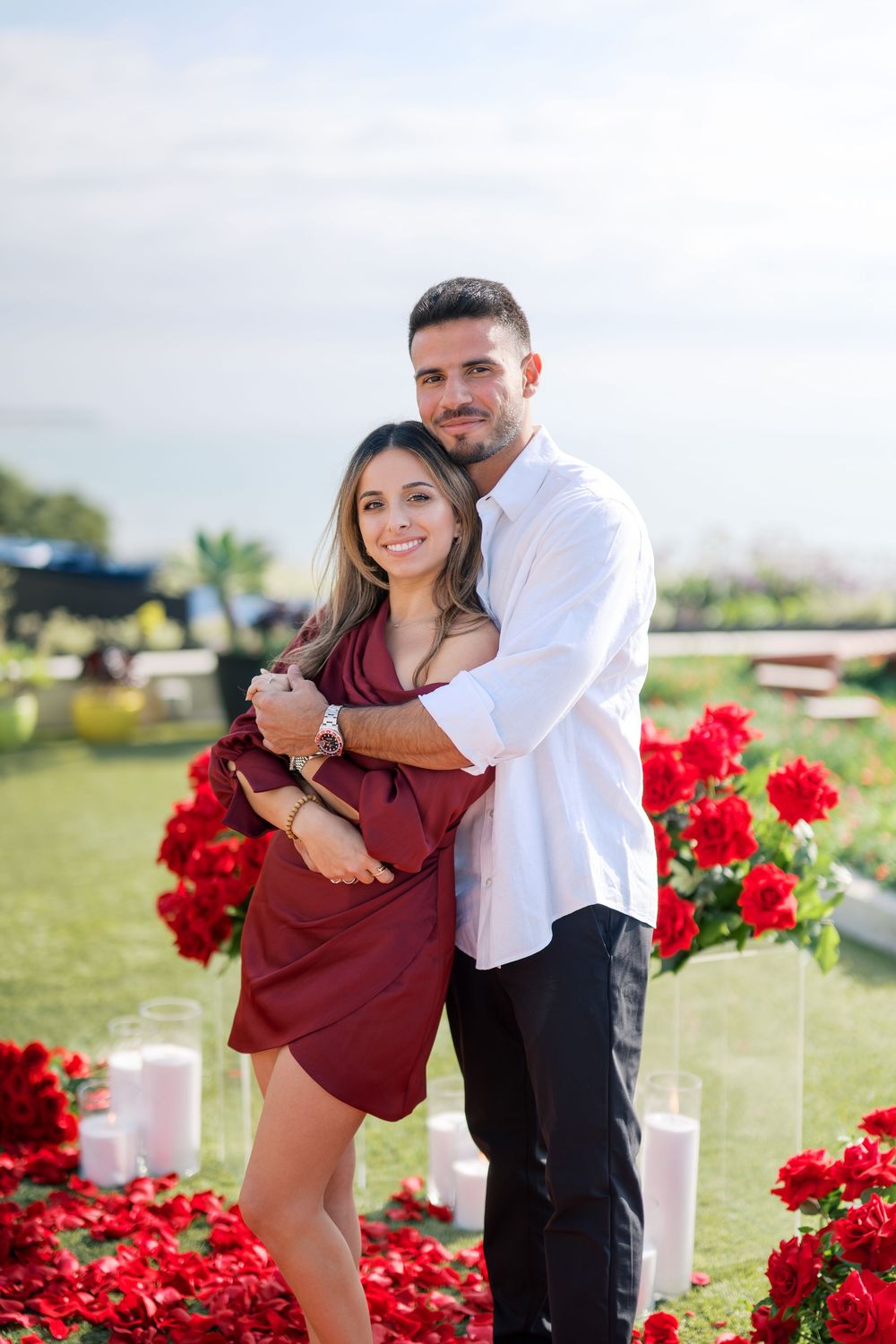 Couple embracing on grass surrounded by red roses and ocean view.