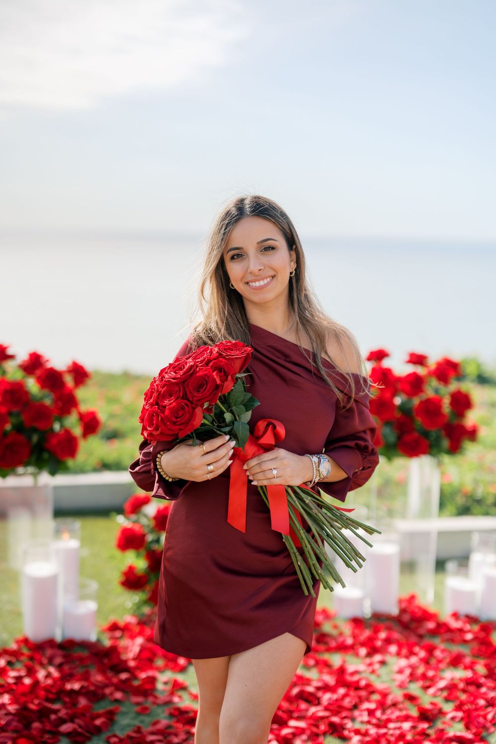 A romantic scene with vibrant red roses and scattered petals along a pathway with a burgundy dress in view.