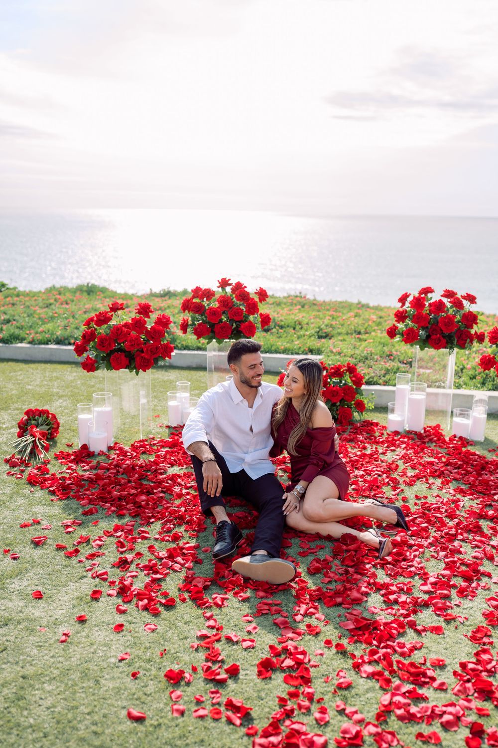 Couple sitting together on grass covered with red rose petals near ocean view.