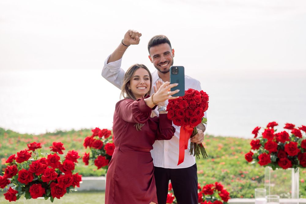 A couple celebrating amidst red roses and scattered petals in a romantic outdoor setting.