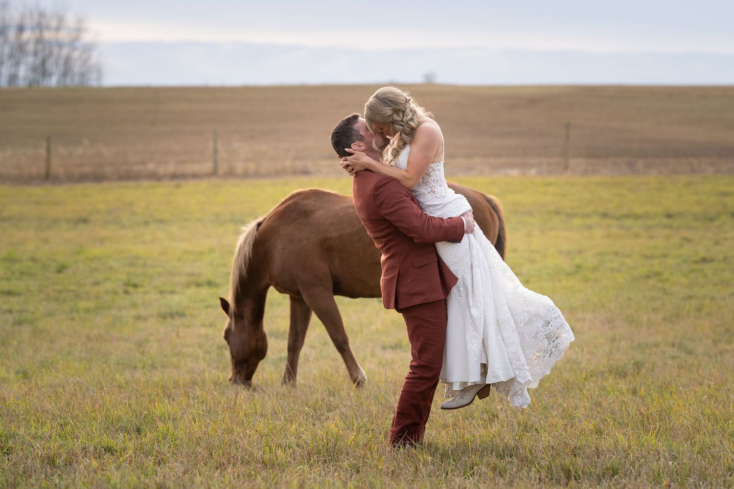 A couple shares a romantic moment in a field while a brown horse grazes peacefully in the background.