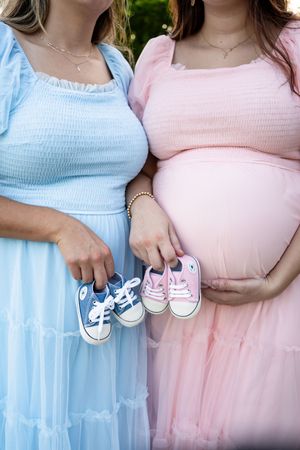 Two expectant mothers in pink and blue dresses holding tiny baby shoes.