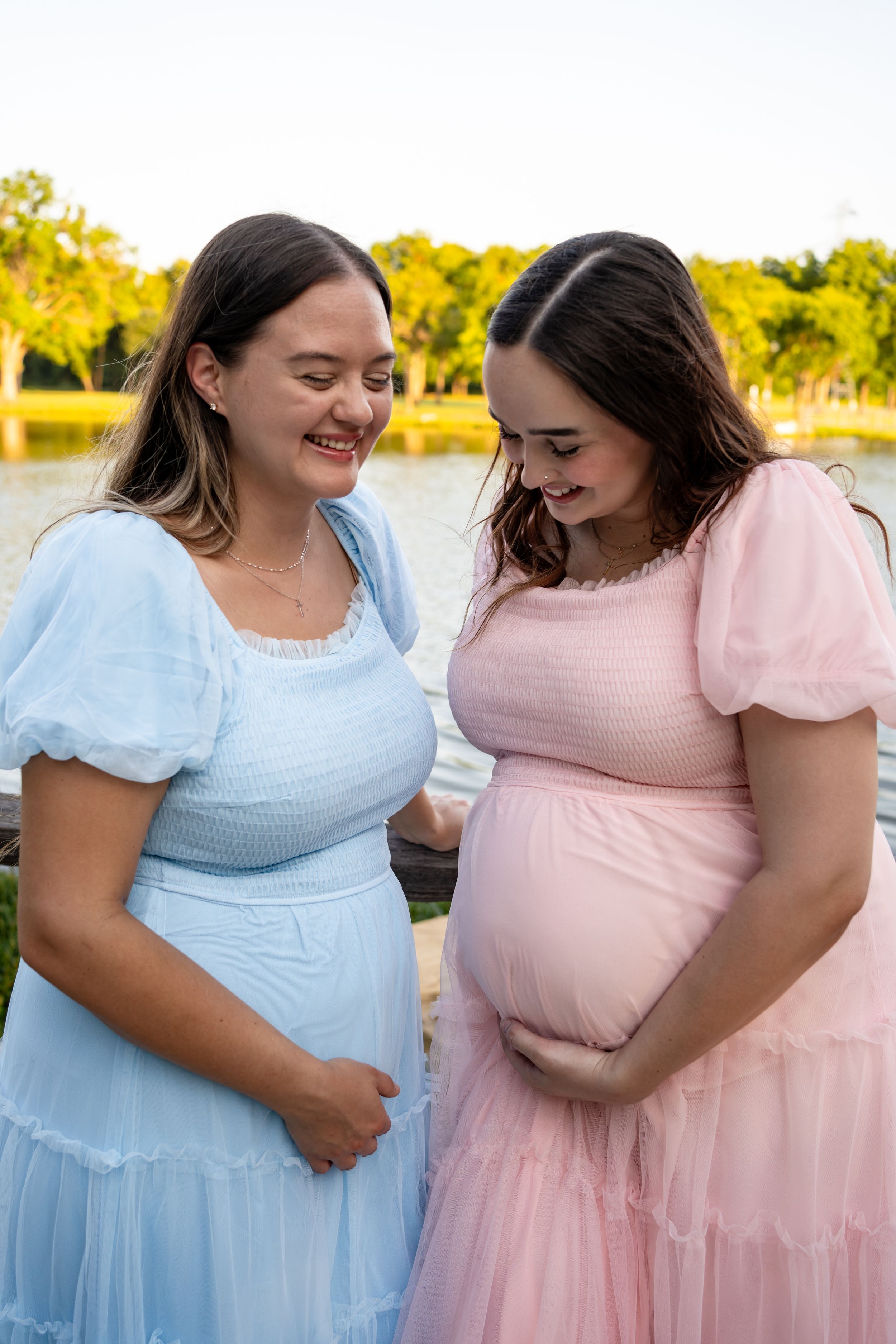Two pregnant women in pastel dresses share a joyful moment by a lake.