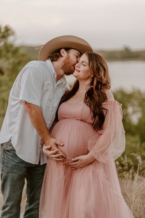 Romantic maternity photoshoot in a field at sunset with couple in pink dress and cowboy hat.