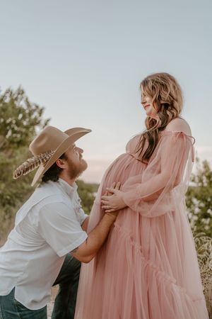 Intimate maternity photos of couple embracing in pink dress and western wear.