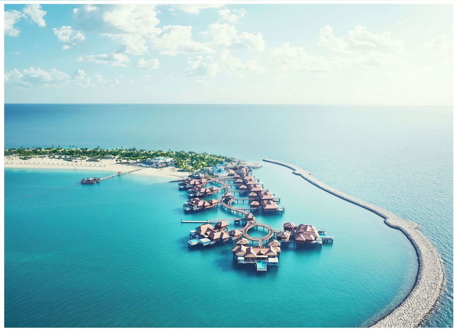 Aerial view of luxury overwater villas at a tropical resort with turquoise waters and curved breakwater jetty.