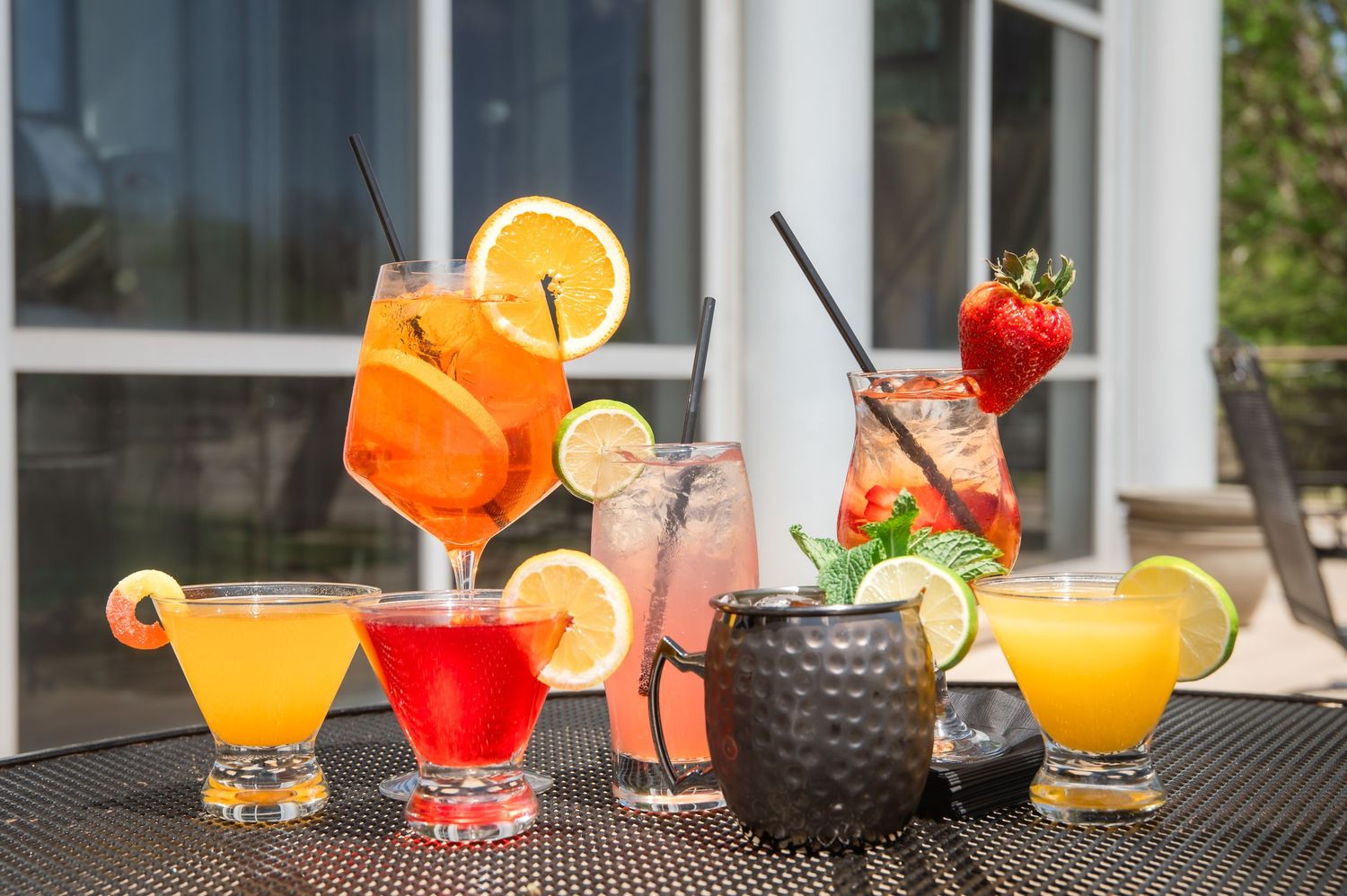 Colorful summer cocktails and mini burgers arranged on a dark bar counter with garnishes and fresh fruit.