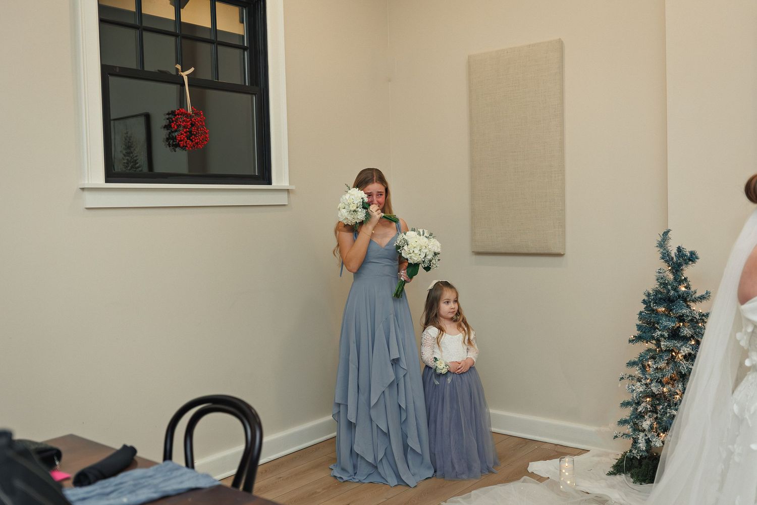 Wedding party members in blue dresses react emotionally in an indoor setting with a Christmas tree.