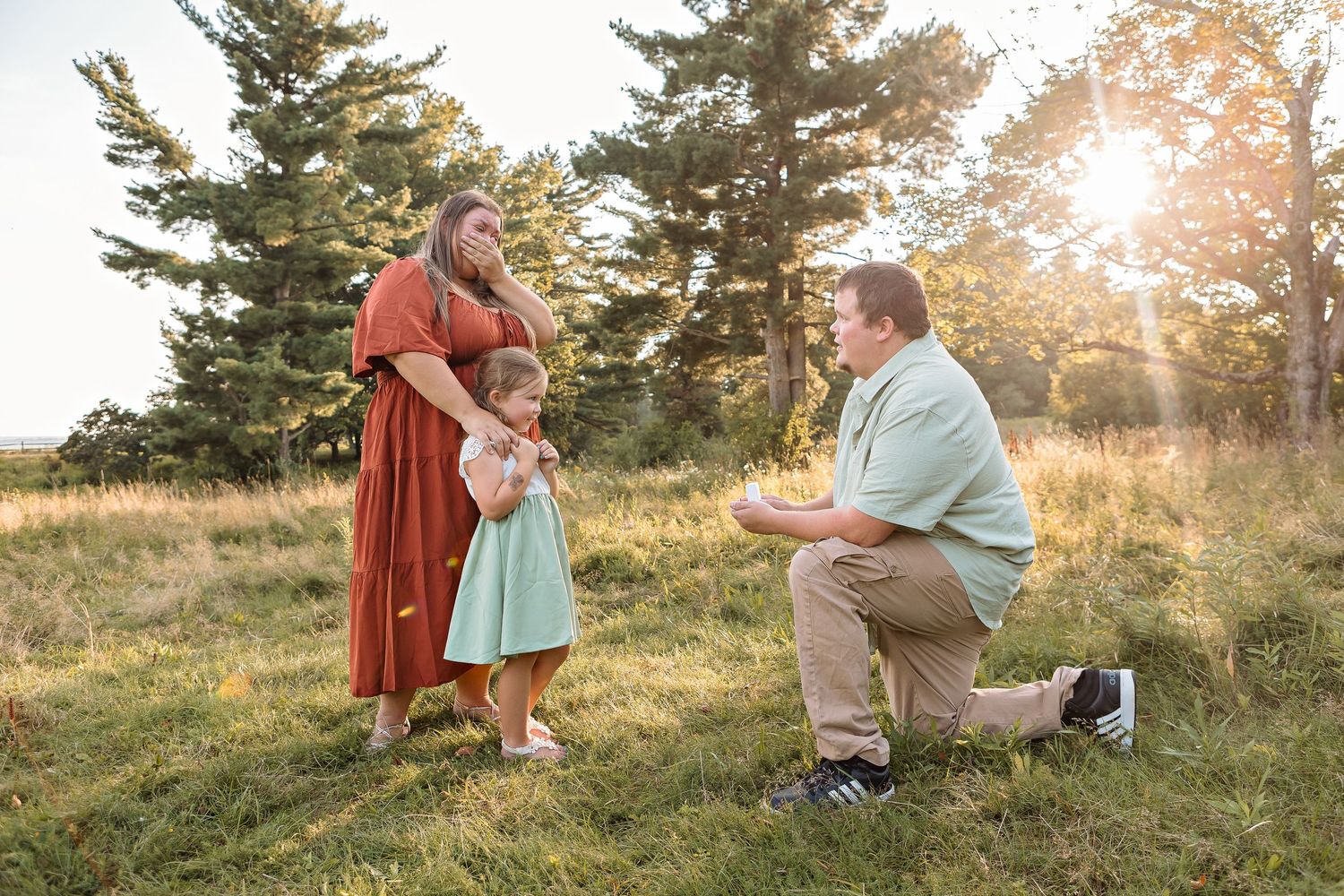 Marriage proposal scene on grassy hillside at sunset with pine trees in background.