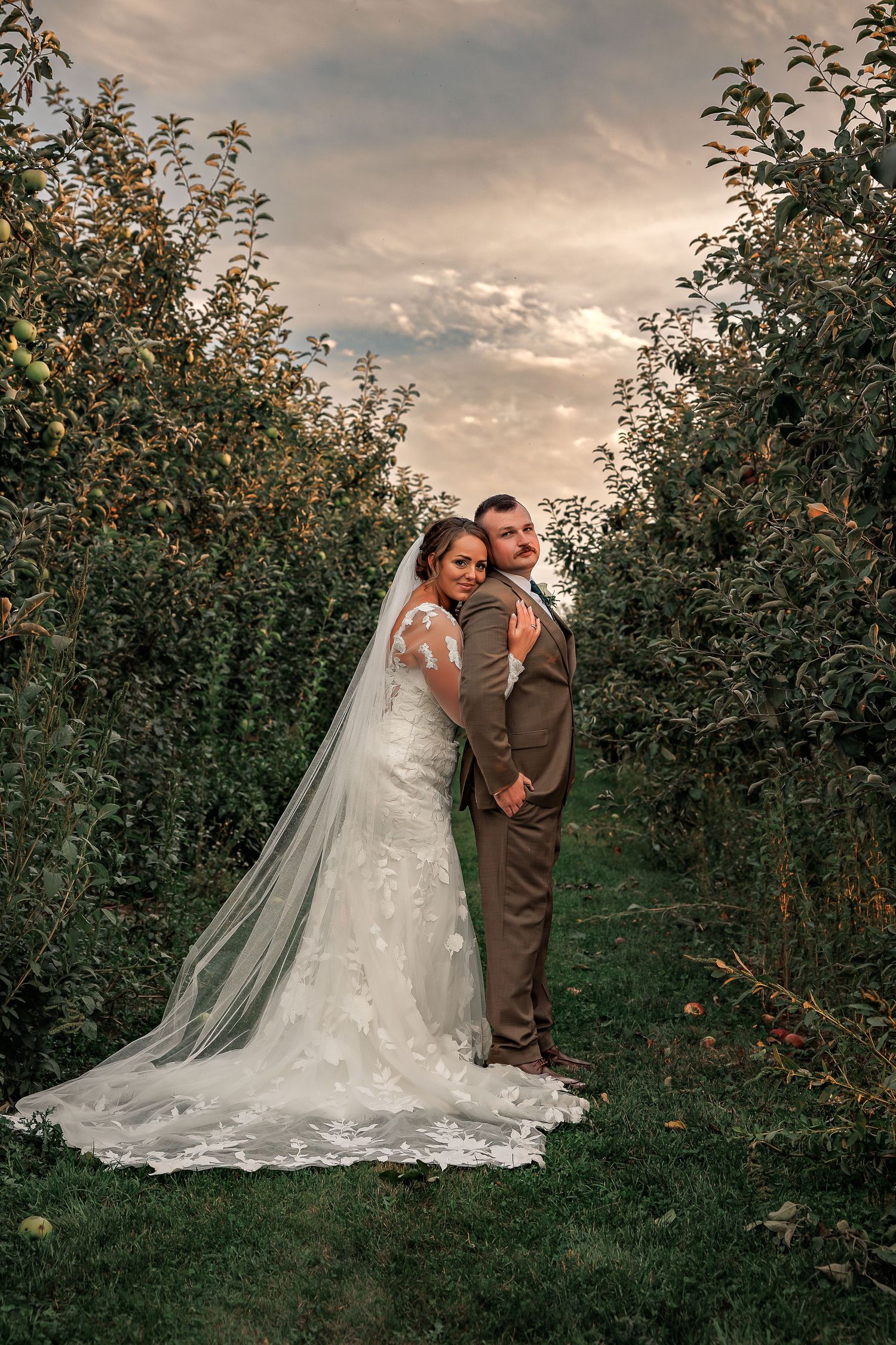 A romantic wedding portrait in an apple orchard at sunset with a flowing white dress and brown suit.