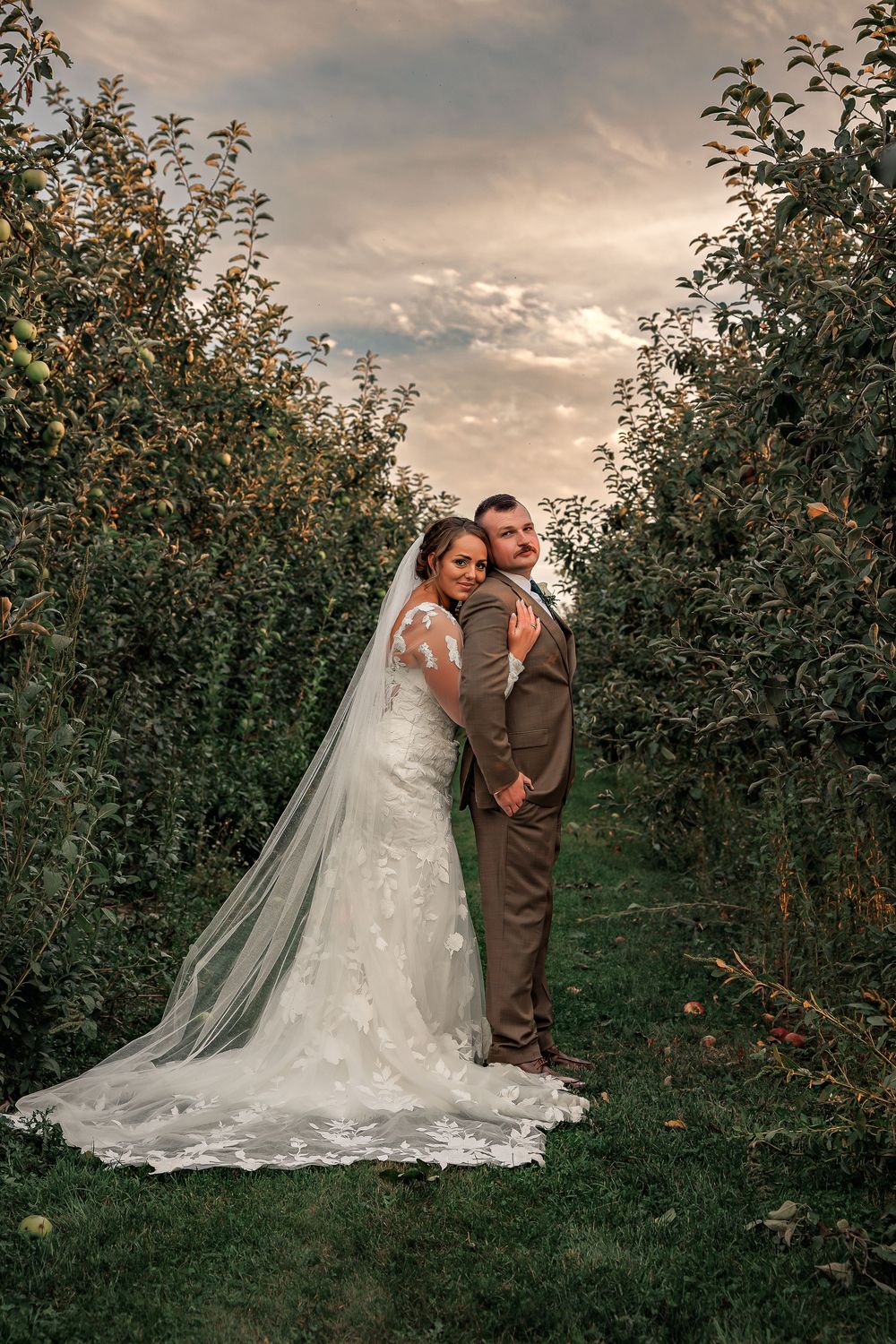 A romantic wedding portrait in an apple orchard at sunset with a flowing white dress and brown suit.