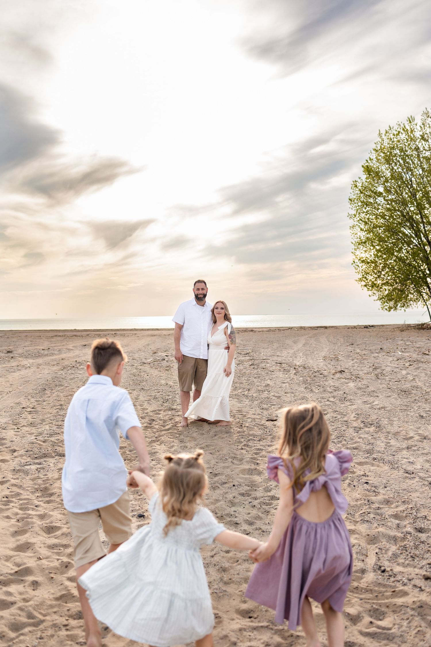 Family plays and runs together on a sandy beach at sunset, creating joyful moments in white and purple outfits.