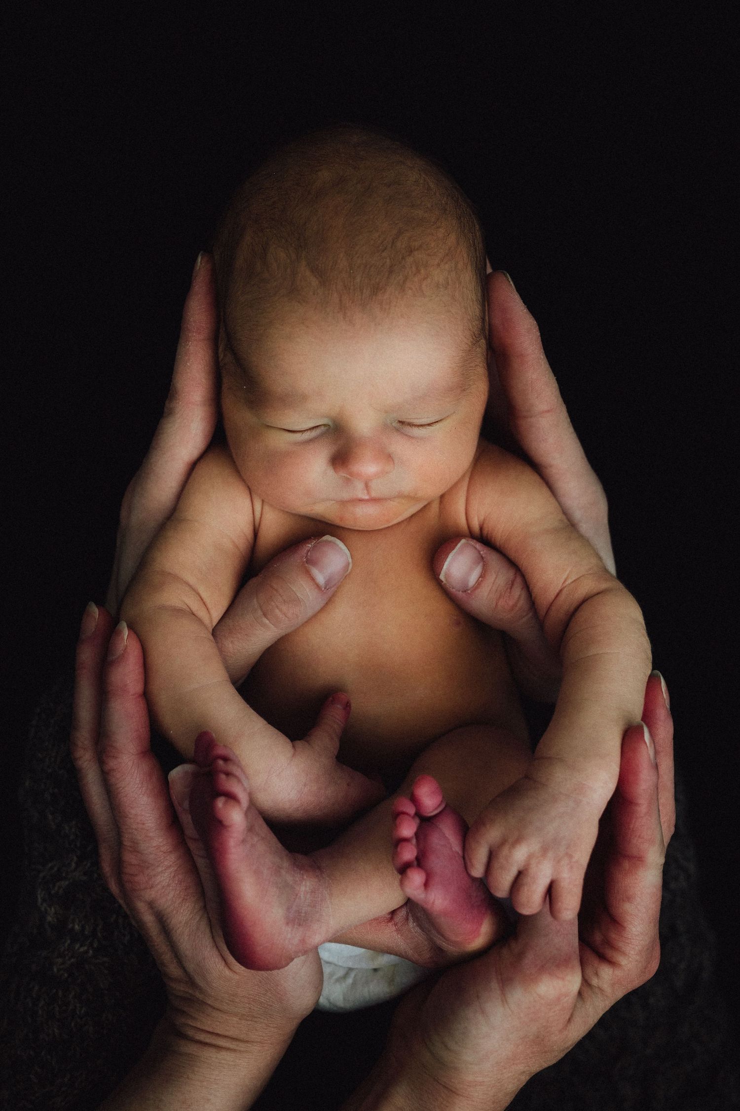 Delicate hands cradle a peaceful sleeping newborn baby against a dark background in an intimate portrait.