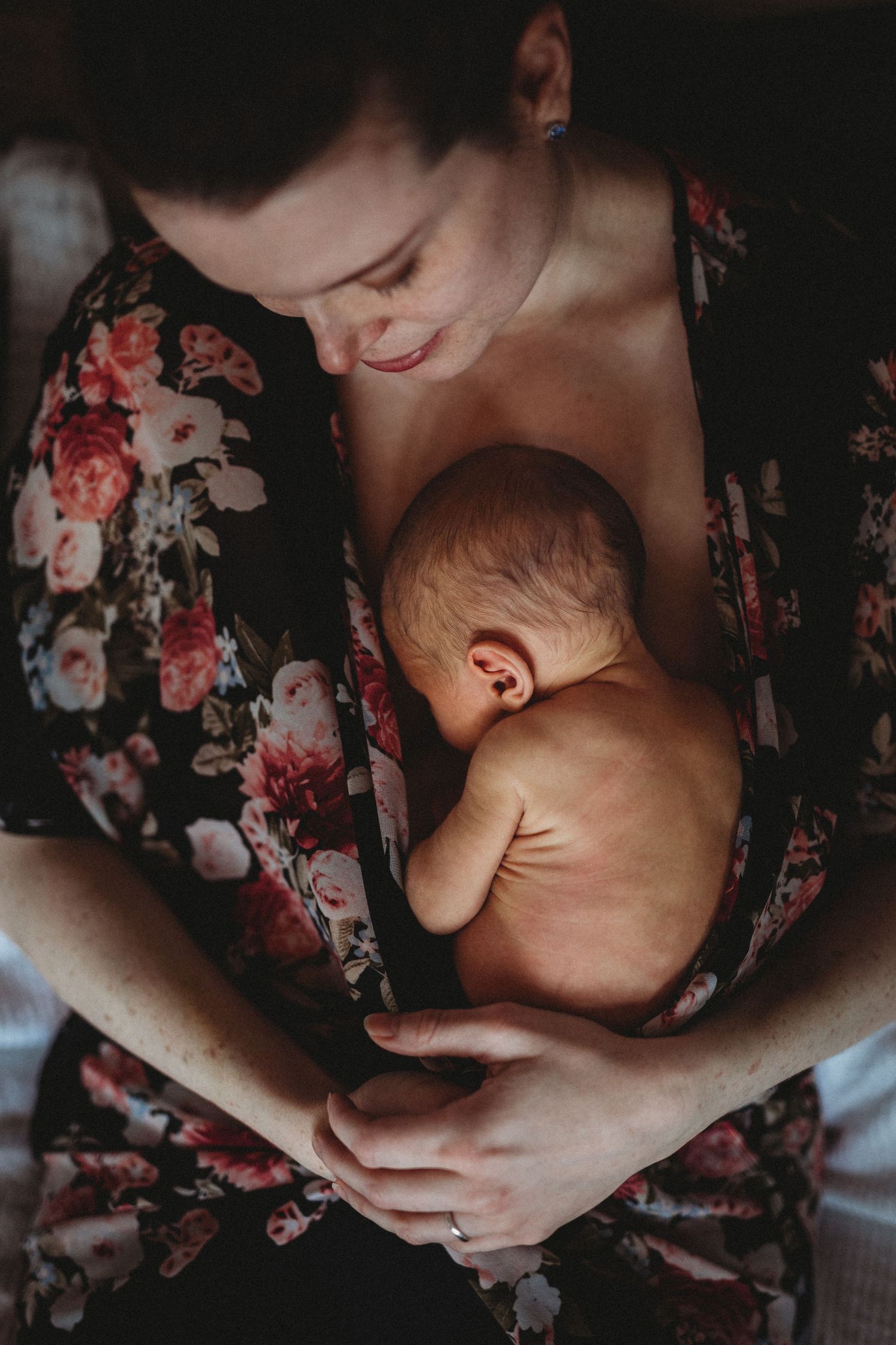 Intimate moody photograph of mother in floral robe holding sleeping newborn baby against her body.