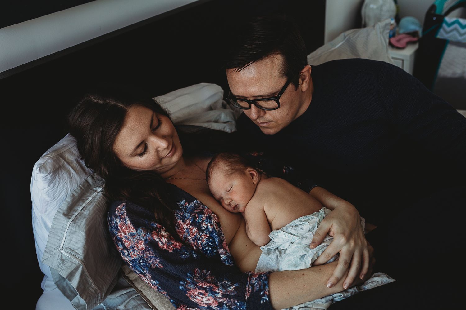 Peaceful family moment as parents lovingly embrace their sleeping baby in moody low light setting.