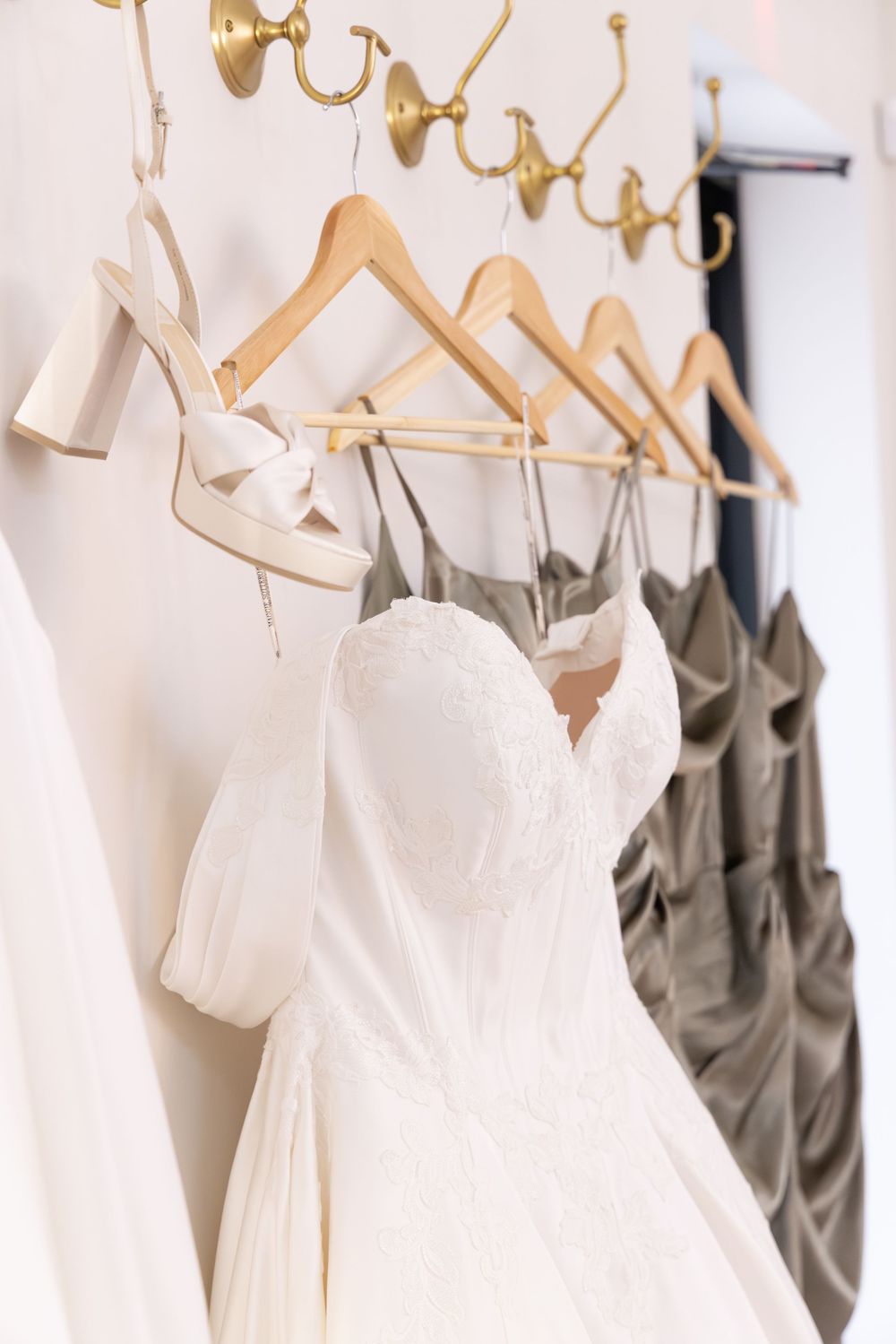 Wedding dresses and gray bridesmaid gowns hanging on golden hangers against a white wall.