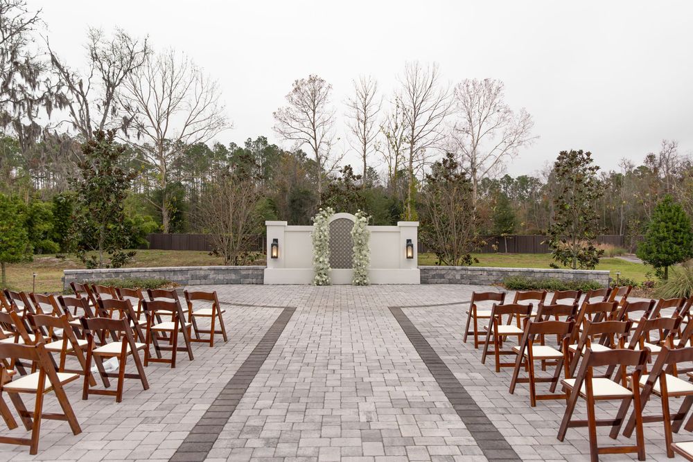 Outdoor wedding ceremony setup with wooden folding chairs and white stone altar on paved courtyard surrounded by trees.