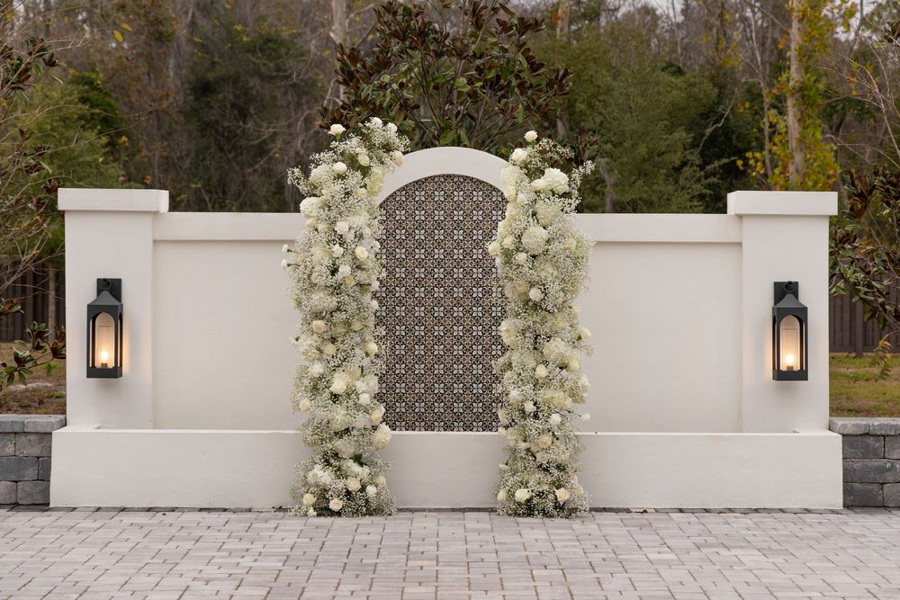 White stone archway with floral arrangements and lanterns decorates an elegant outdoor wedding ceremony space.