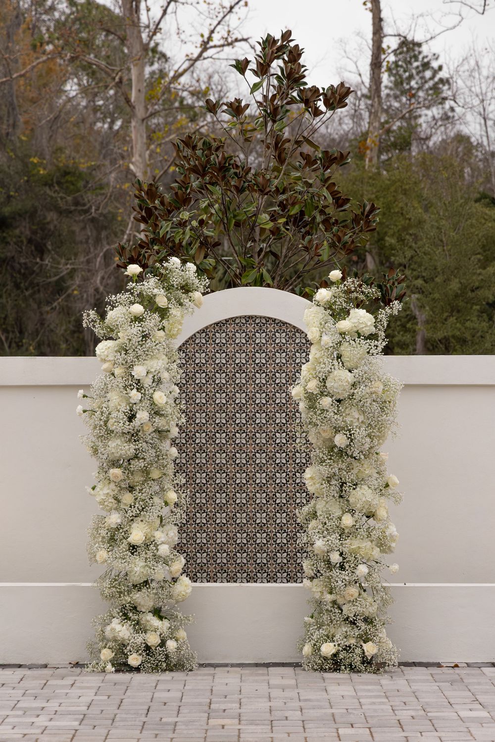 Elegant white floral arch with roses and baby's breath against a cream wall with decorative lattice panel.