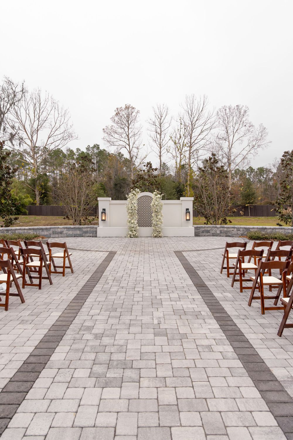 Outdoor wedding ceremony setup with wooden chairs and stone paved aisle leading to a white architectural backdrop.