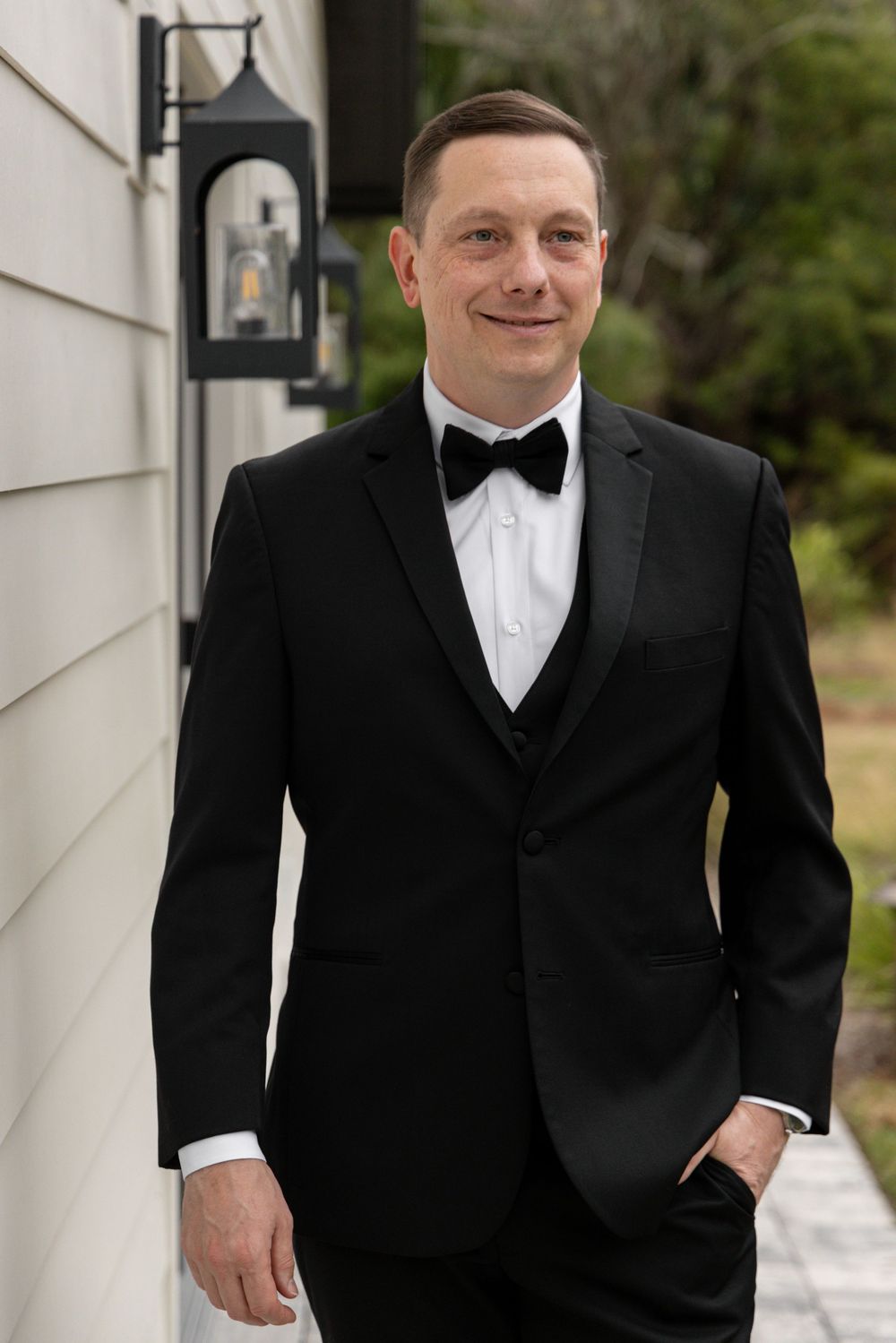 A groom in a classic black tuxedo and white bowtie poses against a light colored wall.