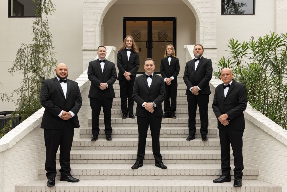 A group of men in formal black tuxedos and bow ties stand on white steps in front of an arched entrance.