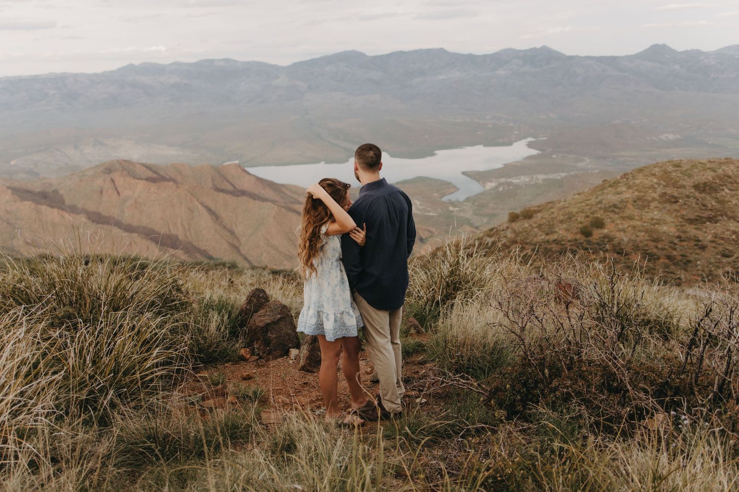 A couple embraces while standing on a scenic mountain overlook with rolling hills and distant landscapes in view.