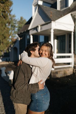 A couple embraces lovingly in front of a white house with dark shutters on a sunny day.