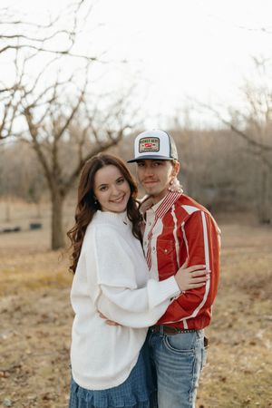 A couple embraces outdoors in a winter field wearing white and red sweaters with blue jeans.