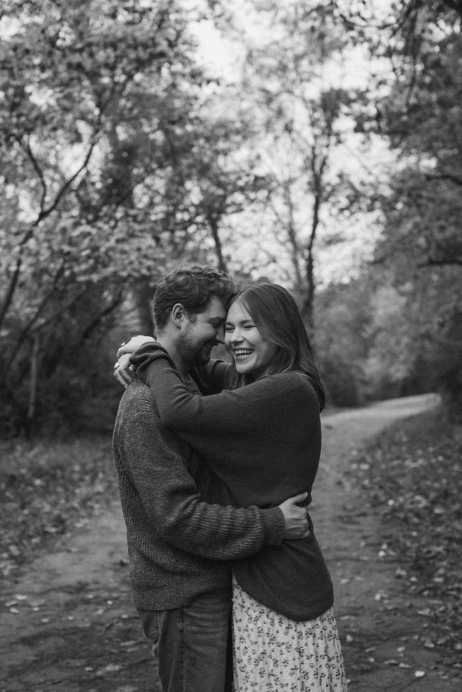 A man and woman embrace and laugh on a tree-lined path during a fall engagement photo session in Kansas City.