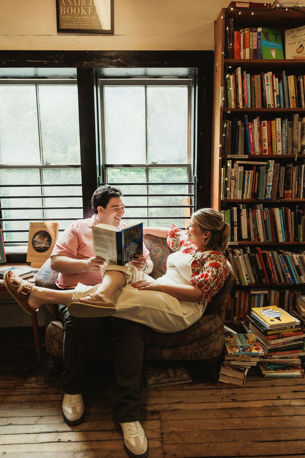 Two people sharing books in a comfortable armchair by a window in a library setting.
