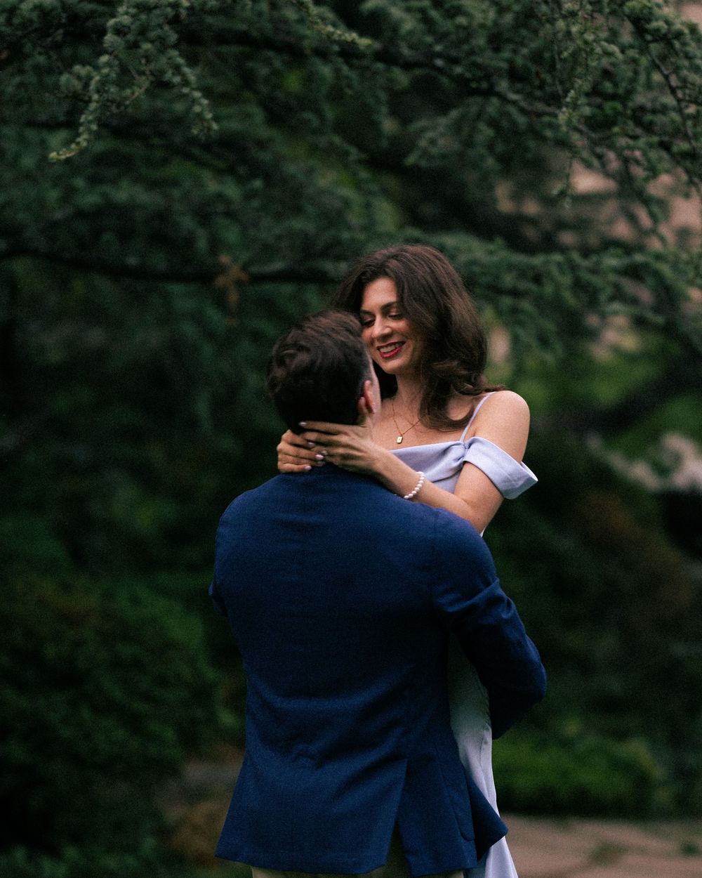 A couple embraces outdoors under lush green trees, woman in off-shoulder dress smiling at man in navy blazer.