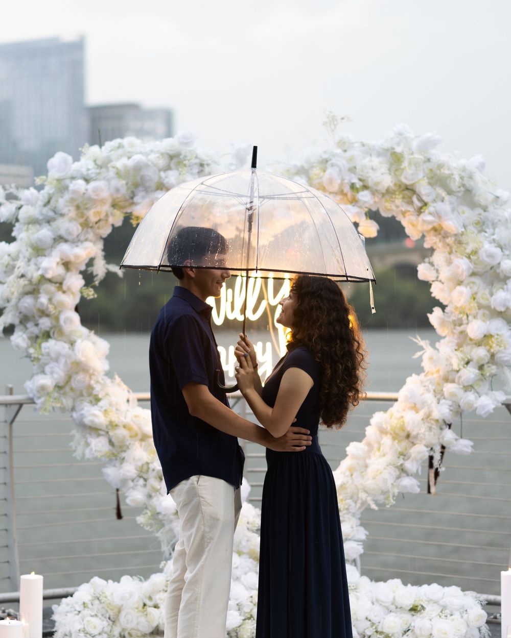 Couple sharing romantic moment under clear umbrella at floral arch wedding proposal setup by waterfront.