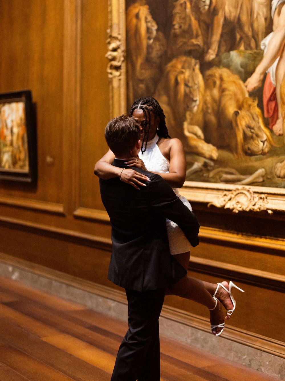 Couple embracing in art museum gallery, man lifting woman in white dress before large classical painting with lions.
