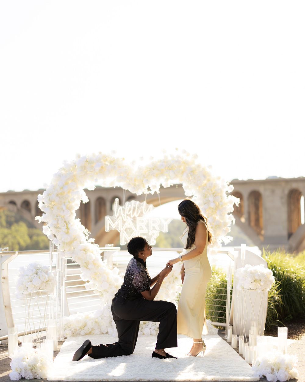 A romantic outdoor proposal with a man on one knee under a white floral heart arch, as a woman in a cream dress accepts.
