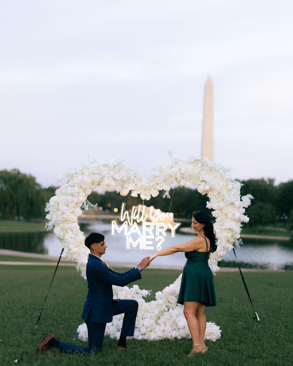 Man proposes to woman with heart-shaped floral arch and Will You Marry Me neon sign near Washington Monument.