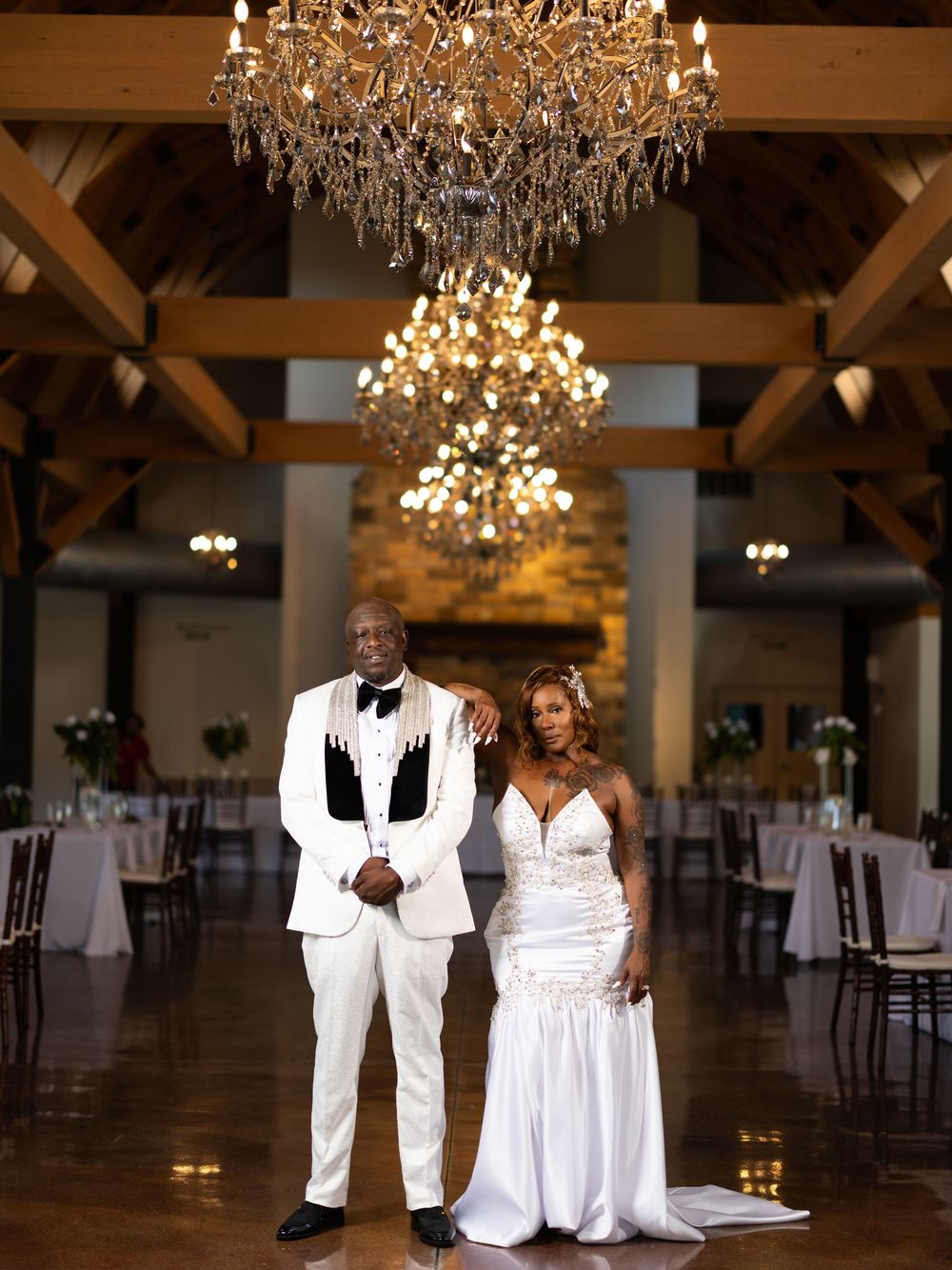 Elegant bride and groom pose under crystal chandelier in rustic wedding venue with exposed beams and reception tables.