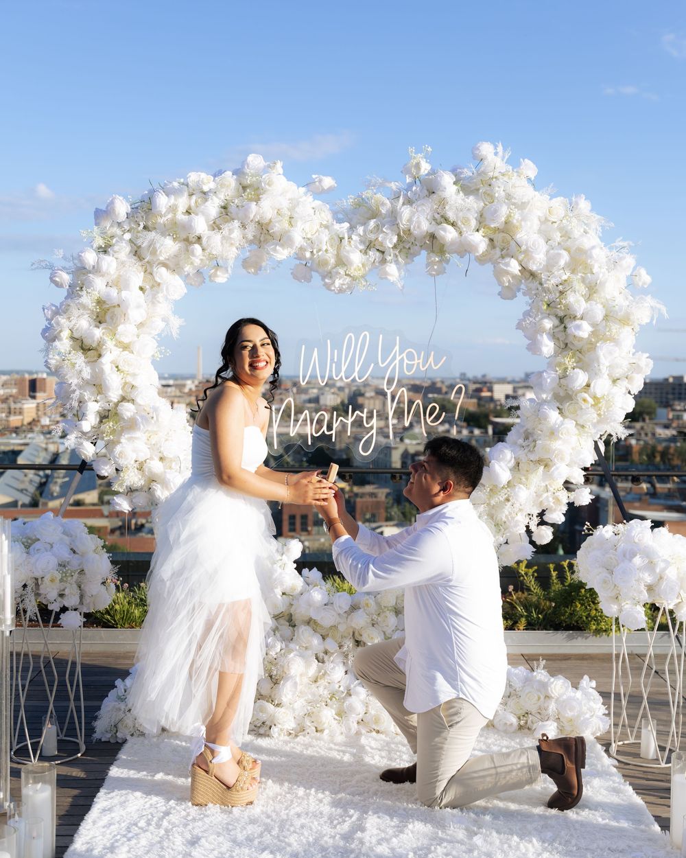 Man proposes to woman under white floral heart arch with Will You Marry Me sign on rooftop.