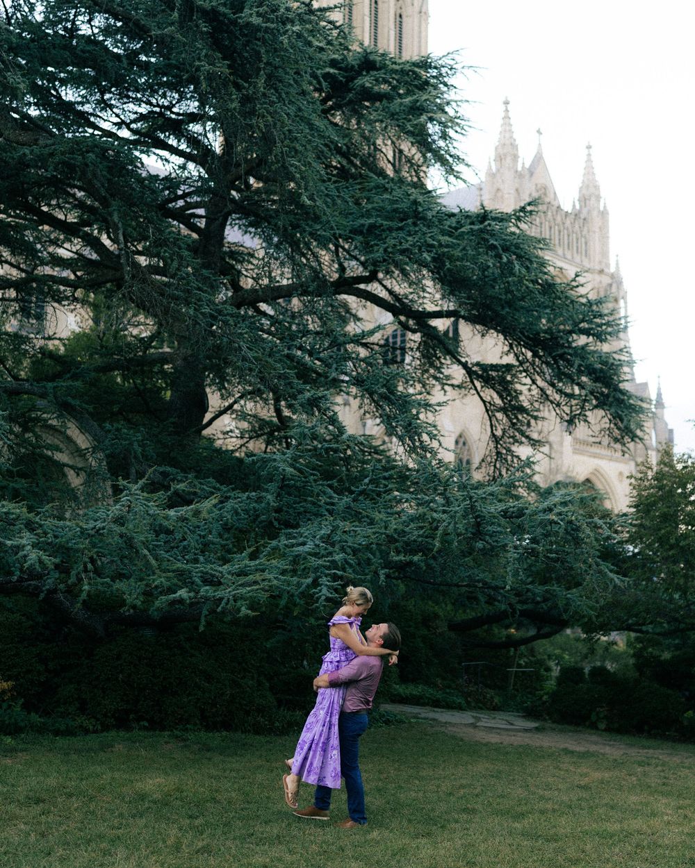 A person lifts a child in a purple dress outdoors near a large cedar tree with a cathedral in the background.