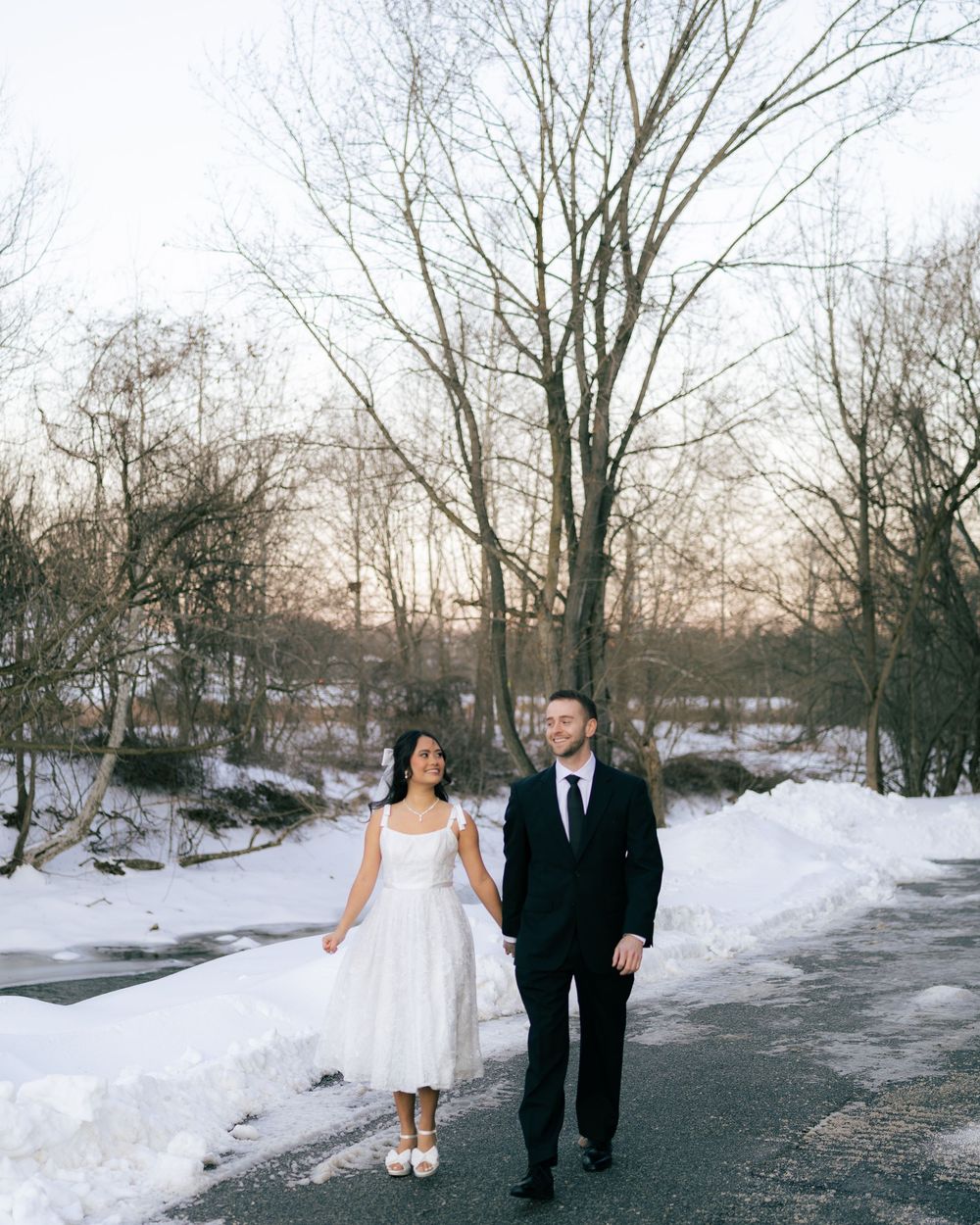 A couple walks along a snowy path through bare winter trees in a romantic outdoor setting.
