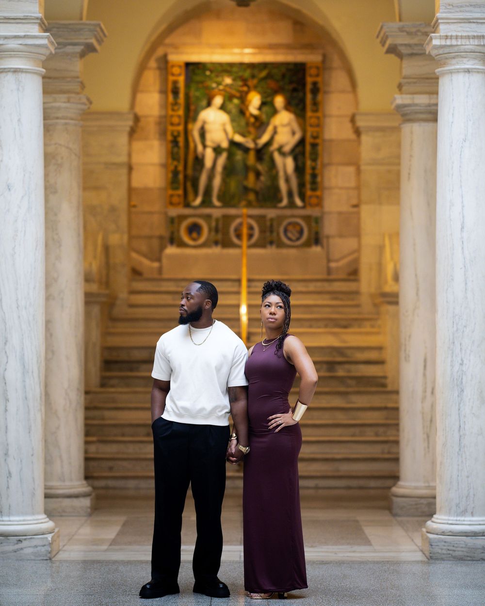 A man and woman pose elegantly in a grand marble hall before a classical painting in an arched alcove.