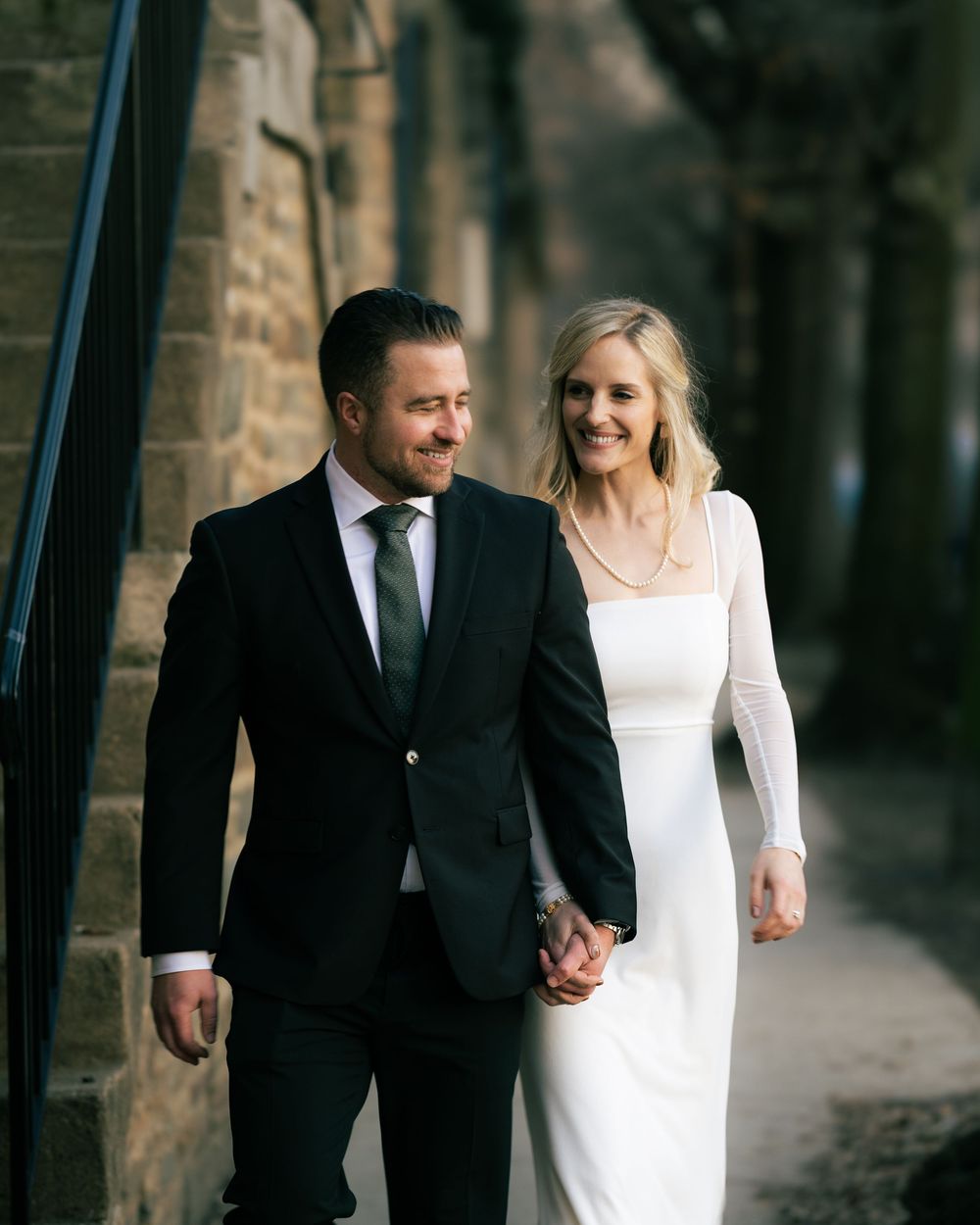A couple in wedding attire walks hand-in-hand, smiling, near a stone building backdrop.