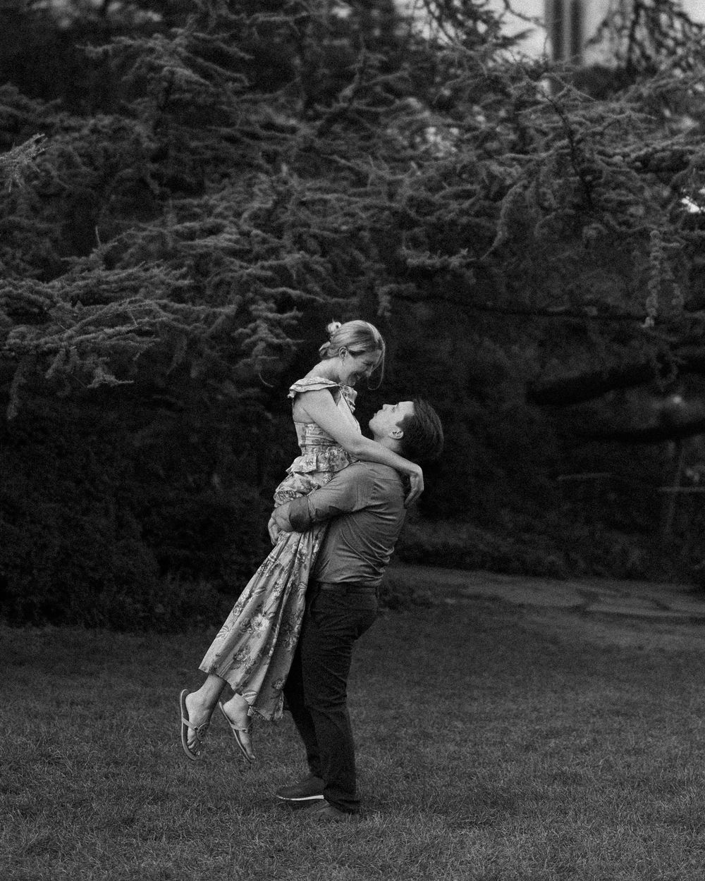 Black and white photo of a man lifting a woman in a floral dress outdoors, with lush trees in the background.